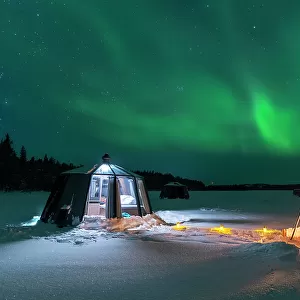 Night view of two people enjoyng dinner outside around a campfire close to the illuminated glass igloo with Northern Lights (Aurora Borealis) dancing in the sky, Jokkmokk, Norrbotten, Swedish Lapland, Sweden, Scandinavia, Europe